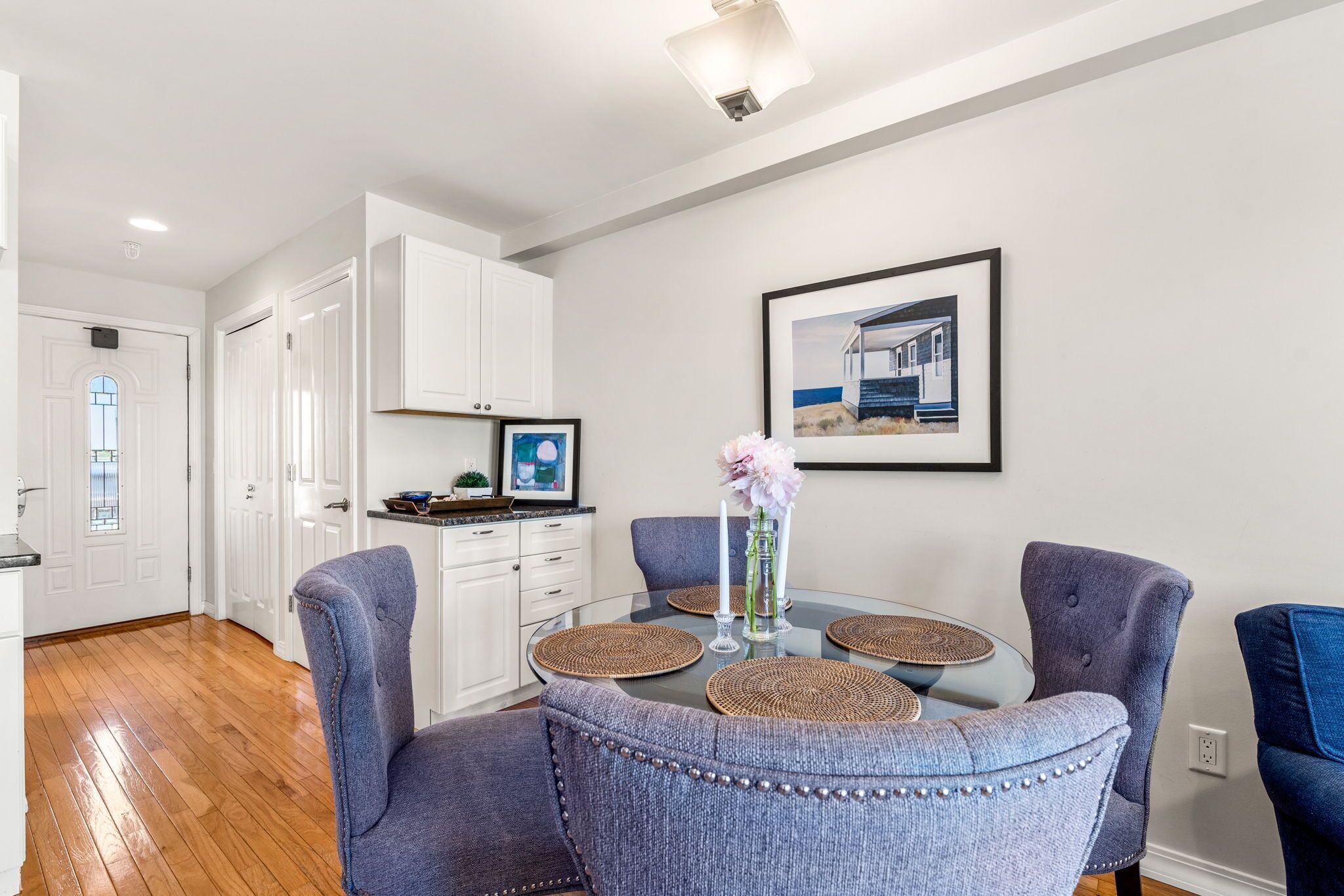 A modern kitchen with white cabinets, a dining area with a glass table and blue upholstered chairs, and a framed picture on the wall.