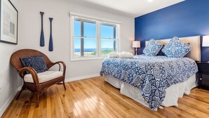 A bedroom with a large bed, a wicker chair, a window with a view, and a blue accent wall.