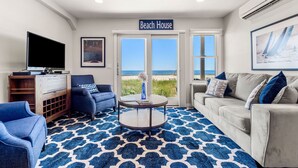 A living room with a large TV, a coffee table, and a sofa. There is a sign that reads "Beach House" and a view of the beach through the windows.