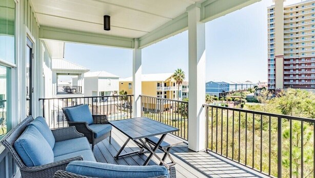 A covered outdoor balcony with wicker furniture and a table, overlooking a residential area with buildings and greenery.