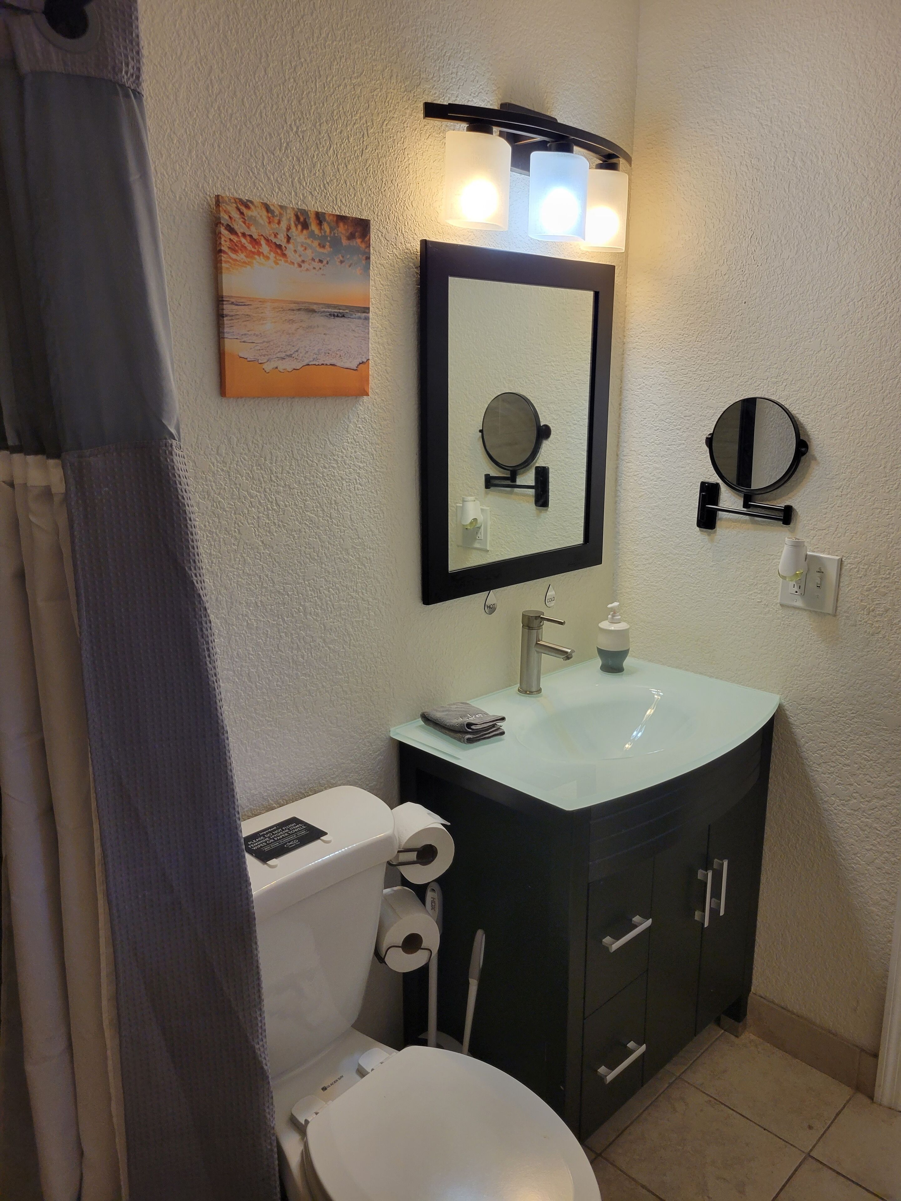 Un baño con inodoro, lavamanos y dos espejos. Hay un perchero y un cuadro en la pared.