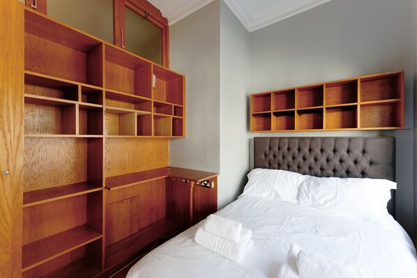 A bedroom with a bed, a wooden wardrobe, and a built-in shelf above the bed.