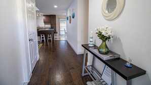 A hallway with a dark wood floor, a white door, a small table with a vase of flowers, and a mirror on the wall.