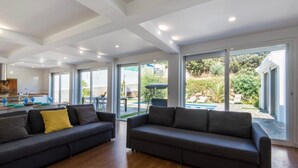 A spacious living room with two dark sofas, a wooden floor, and large sliding glass doors leading to an outdoor area with a pool and greenery. - Pool Villa & Beach House (Torre de Benagalbón)