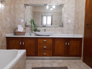 Un bany amb una banyera blanca, un mirall gran, un lavabo blanc, armaris de fusta i una petita planta.