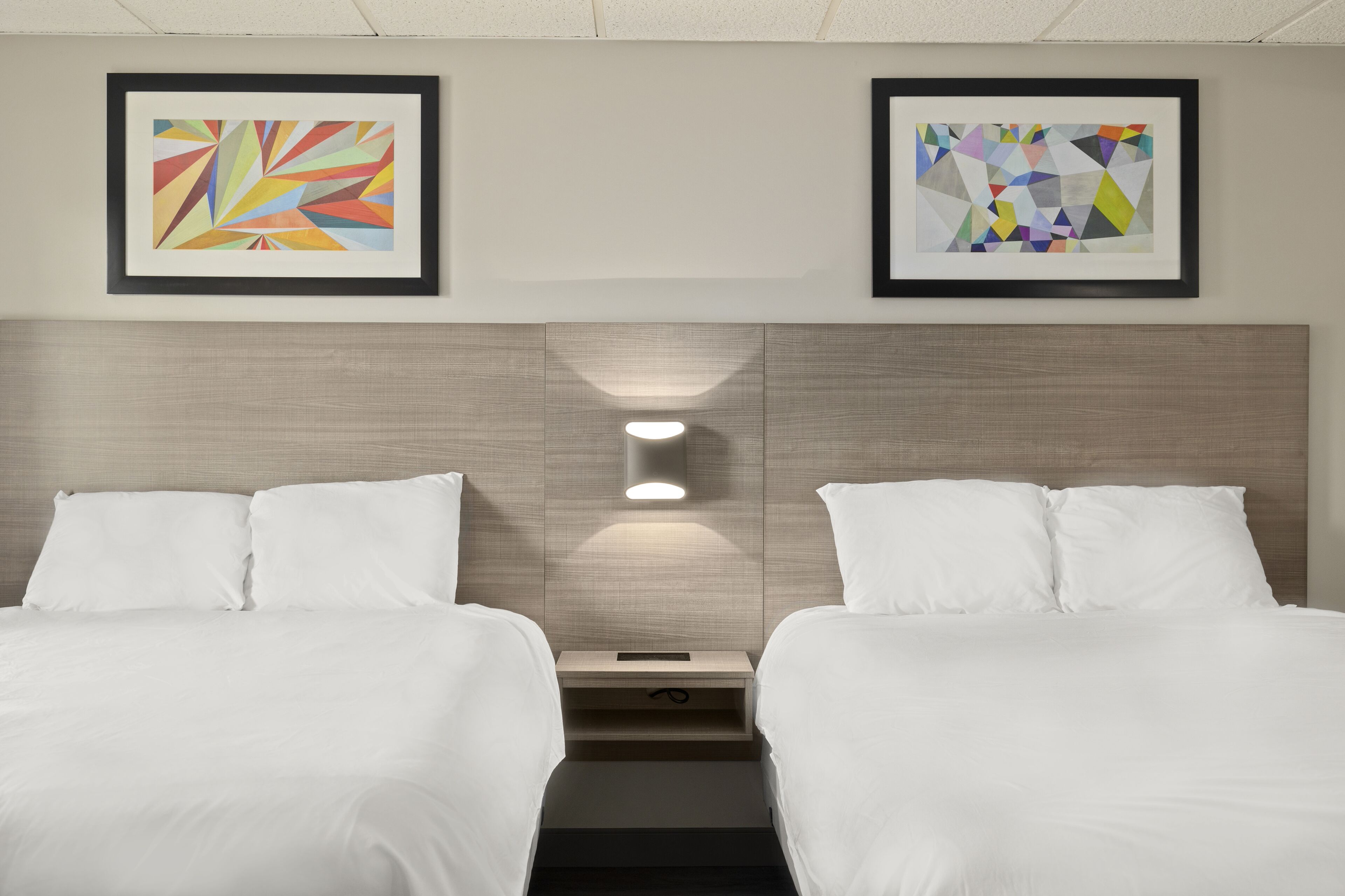 A hotel room with two beds, a nightstand, and two framed abstract artworks on the wall.