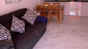 Uma sala de estar moderna com um sofá cinza escuro, almofadas estampadas, uma mesa de jantar com cadeiras e uma cozinha com armários brancos e micro-ondas.