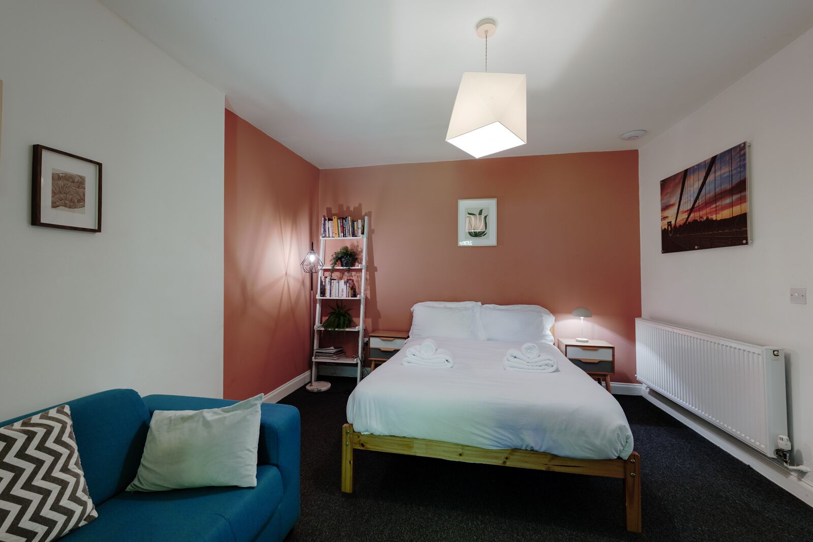 Un dormitorio con una cama, un sofá azul, una estantería, un radiador y un cuadro en la pared.
