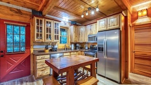 A wooden cabin interior with a kitchen featuring wooden cabinets, a stainless steel refrigerator, and a wooden dining table with stools. - Twin Creeks Refuge, a log cabin, Pet friendly (Maggie Valley)