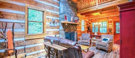 A log cabin interior with a stone fireplace, wooden furniture, and a balcony.