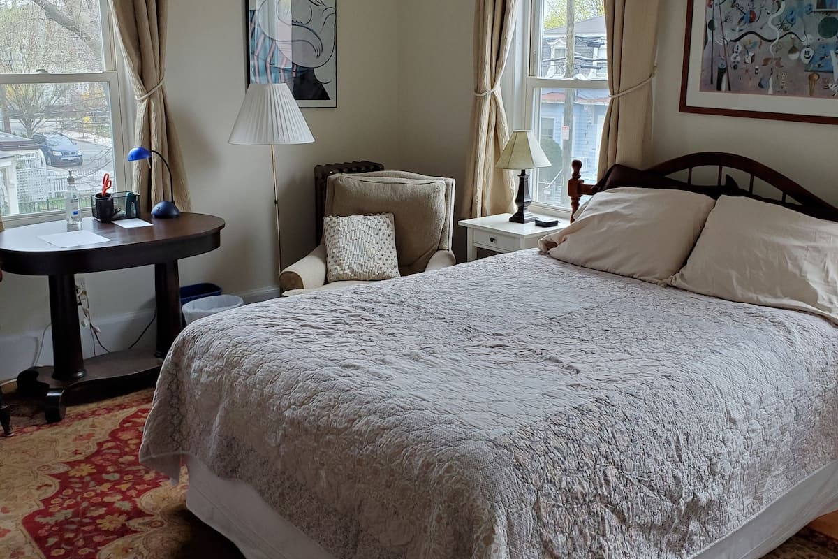 A neatly arranged bedroom with a large bed, a desk, and a chair. There are two windows with curtains, a lamp on the desk, and a painting on the wall.