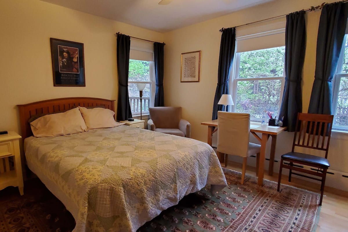 A hotel room with a bed, a chair, a desk, and two windows with curtains.