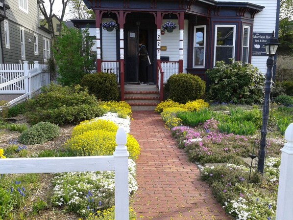 A traditional house with a front porch, a brick pathway, and a variety of flowering plants.