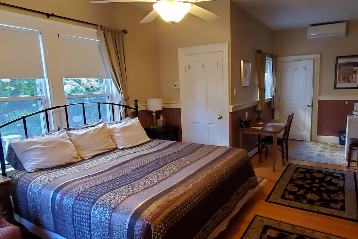 A bedroom with a bed, bedside table, lamp, and a desk with a chair. There is a door and a window with curtains.