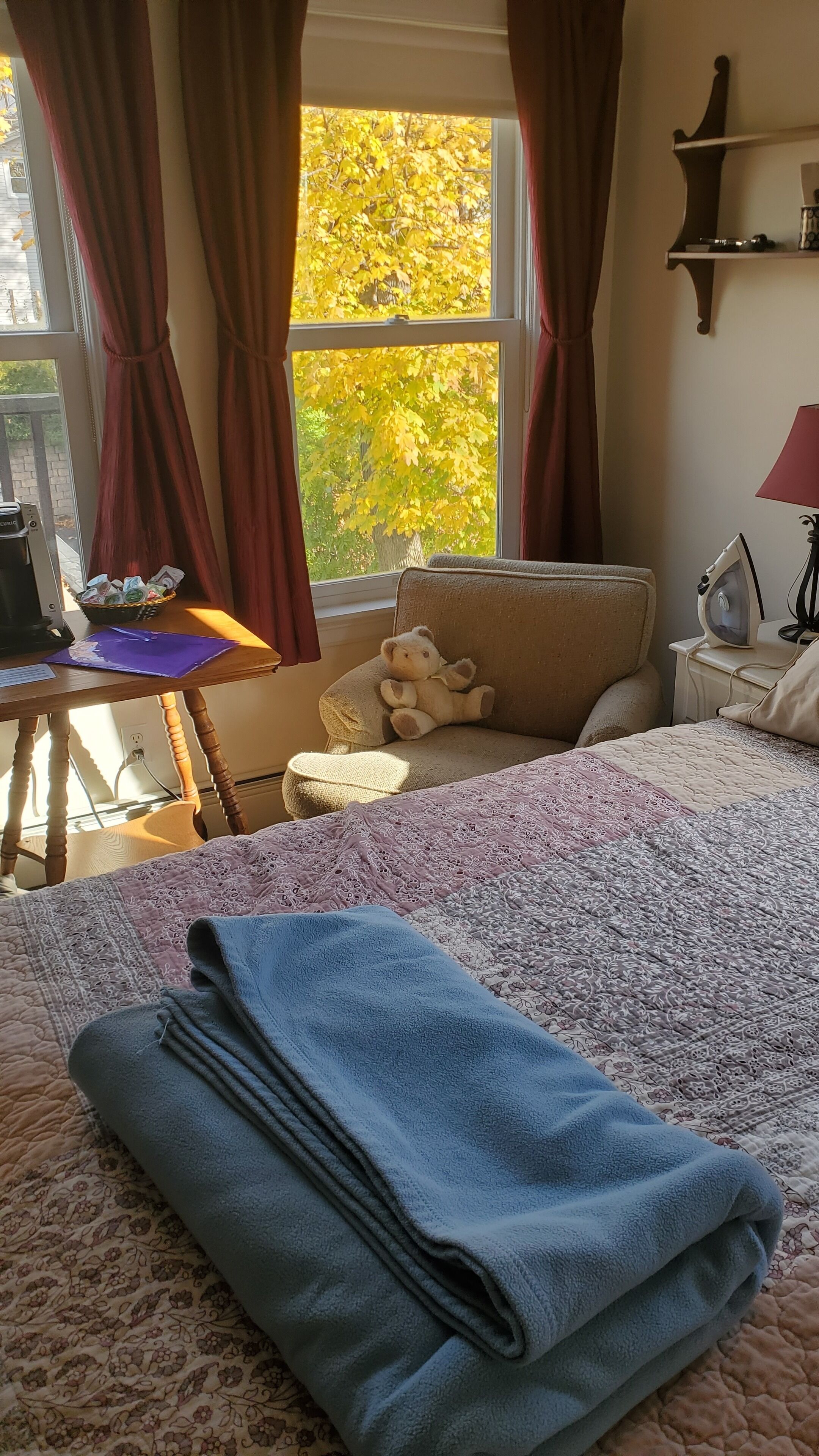 A bedroom with a bed, a chair, a window with curtains, a lamp, and a teddy bear on the chair.