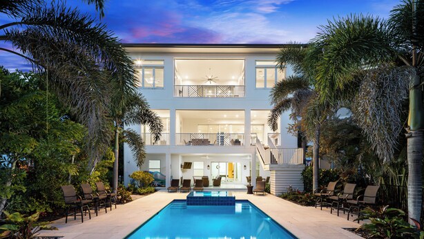 A two-story building with a swimming pool in the foreground, surrounded by palm trees and outdoor seating.