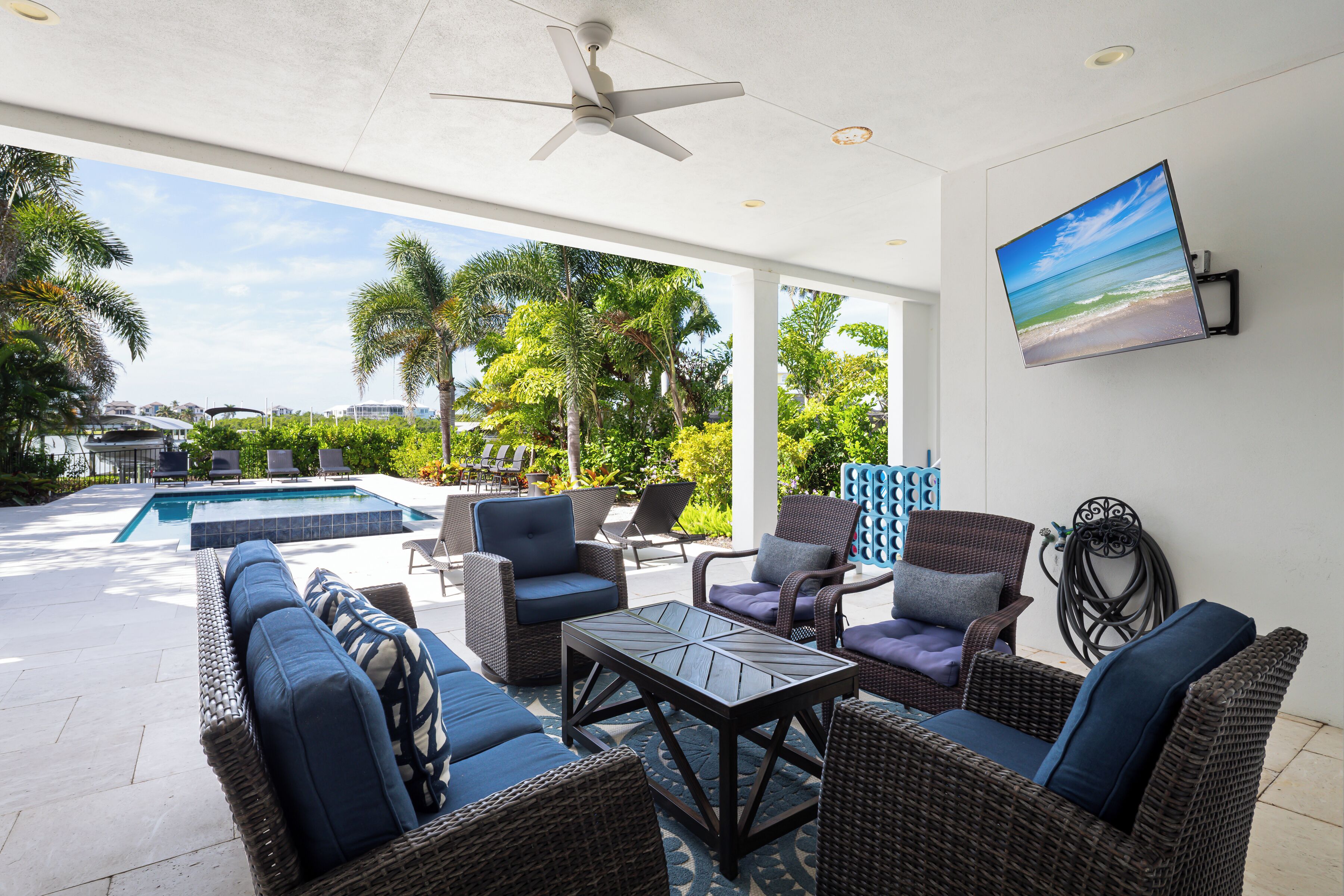 A covered outdoor patio with wicker furniture, a ceiling fan, and a view of a swimming pool and palm trees.