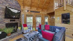 A cozy cabin interior with a stone fireplace, a television mounted on the wall, a blue sofa with colorful cushions, and a wooden ceiling. - Incredible Vibes & Views #204 (Sevierville)