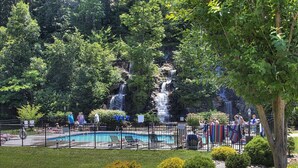 A swimming pool surrounded by a fence, with a waterfall in the background and well-maintained bushes in the foreground. - Incredible Vibes & Views #204 (Sevierville)