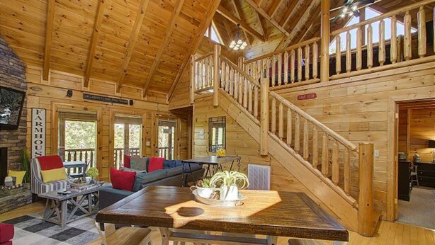 A spacious log cabin with a high ceiling, a central dining area with a wooden table and chairs, a staircase leading to an upper level, and a cozy living room with a sofa and a fireplace. - Incredible Vibes & Views #204 (Sevierville)