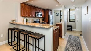 Una cocina moderna con encimeras de granito, armarios de madera y un microondas integrado. Hay tres taburetes de bar y una pequeña isla con un fregadero.