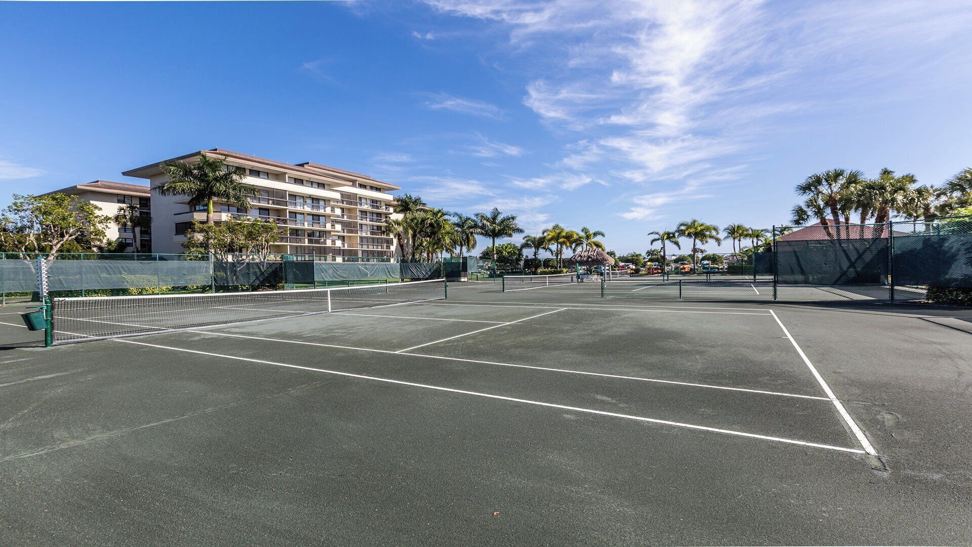 Un court de tennis en plein air, avec un filet, entouré de palmiers et avec un bâtiment d’hôtel en arrière-plan.
