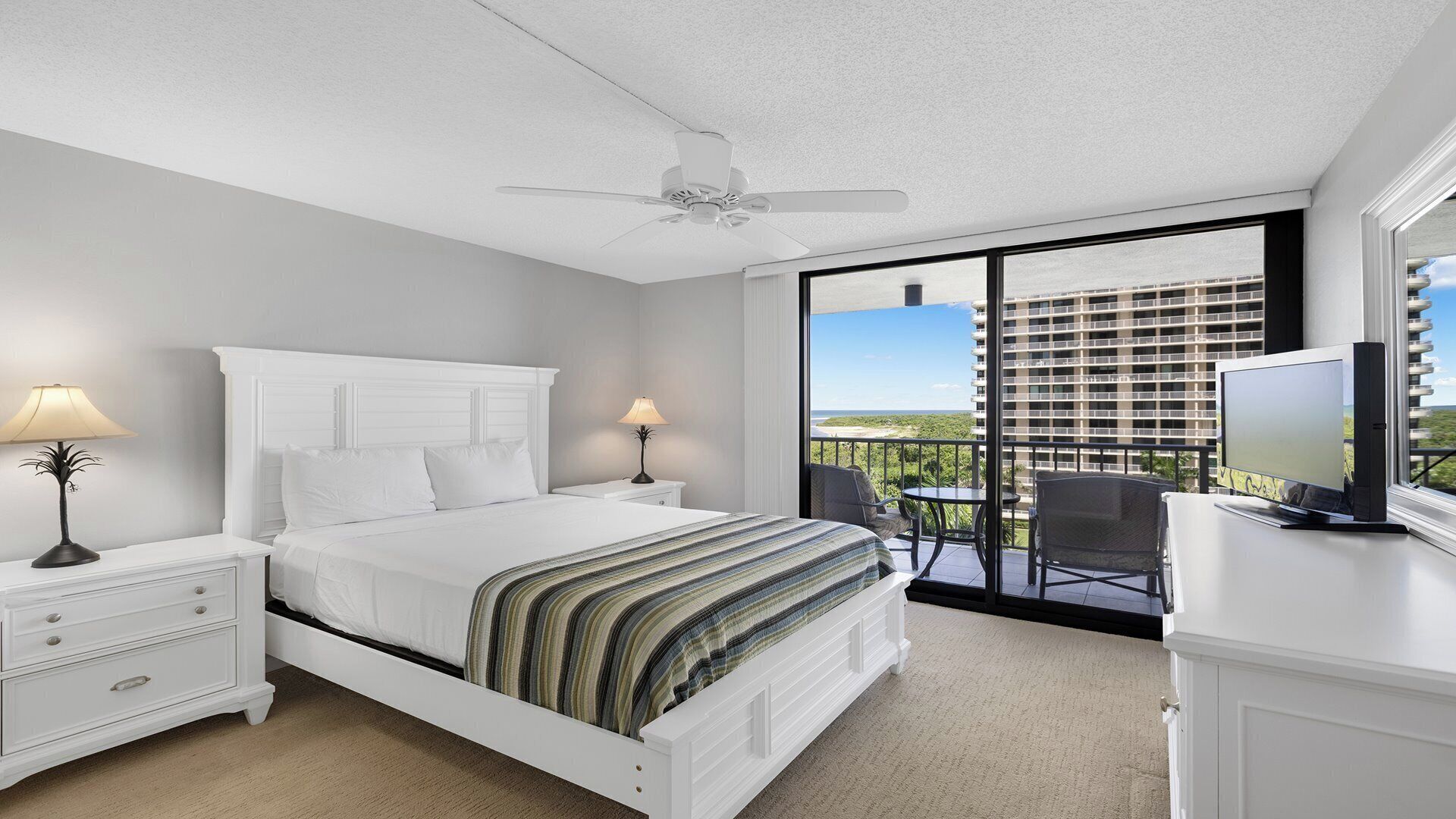 A hotel room with a bed, two bedside lamps, a ceiling fan, a balcony with a TV, and a view of a building.