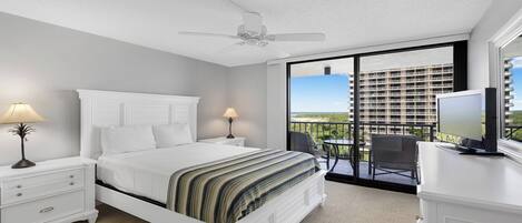 A hotel room with a bed, two bedside lamps, a ceiling fan, a balcony with a TV, and a view of a building.