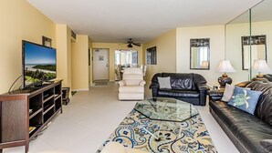 A living room with a TV, leather sofas, a glass coffee table, and a hallway leading to a dining area. - This 2/2 condo is just steps away from the beach (Marco Island)