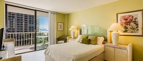 Quarto de hotel com uma cama grande, mesa de cabeceira, cadeira, escrivaninha e vista para a cidade através de uma varanda.