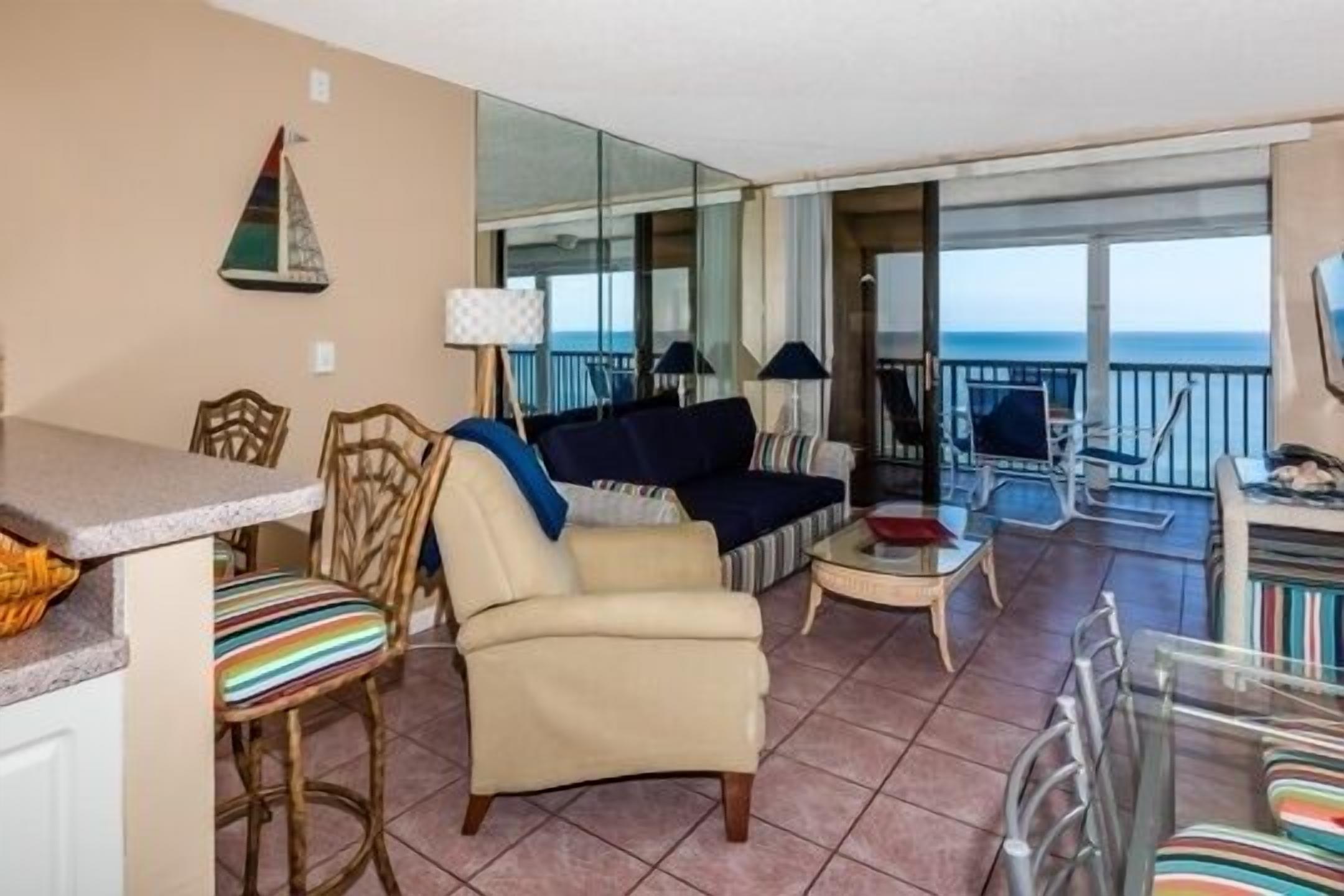A hotel room with a balcony, a sofa, a chair, a small table, and a view of the sea.