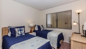 Une chambre avec deux lits, chacun recouvert d’une couvre-lit bleue et agrémenté d’oreillers assortis, une table de chevet avec une lampe, et une fenêtre avec des stores.