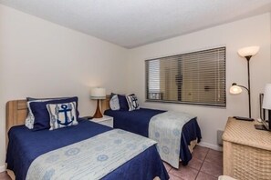 A room with two beds, each with a blue bedspread and matching pillows, a bedside table with a lamp, and a window with blinds.