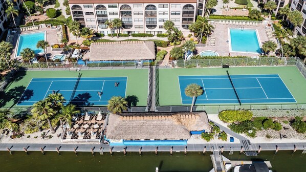 An aerial view of a resort with two tennis courts, a pool area, and a building with multiple balconies.