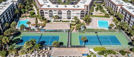 Vista aérea de un hotel con dos canchas de tenis, piscina y zona de playa con sombrillas.