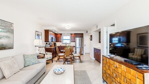 A modern living room with a sofa, a dining area with chairs, a kitchen with cabinets and appliances, and a TV on a wooden cabinet.