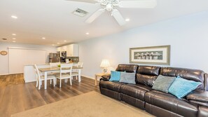 A modern living room with a leather sofa, a dining area with a white table and chairs, a kitchen with white cabinets, and a ceiling fan.