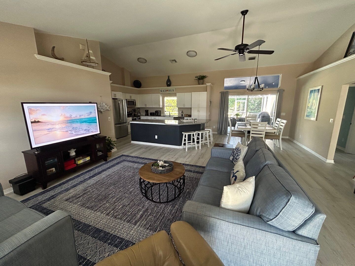 A spacious living room with a large gray sofa, a coffee table, and a flat-screen TV. There is a kitchen area with white cabinets and a central island.