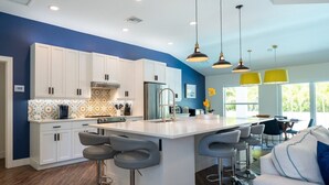 A modern kitchen with white cabinets, a central island with bar stools, and a dining area with yellow pendant lights.