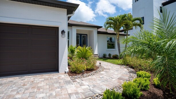 A modern two-story house with a garage, a stone-paved driveway, and a well-maintained garden with various plants and a palm tree.