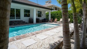 A swimming pool with a white house in the background, surrounded by palm trees and a clear sky.