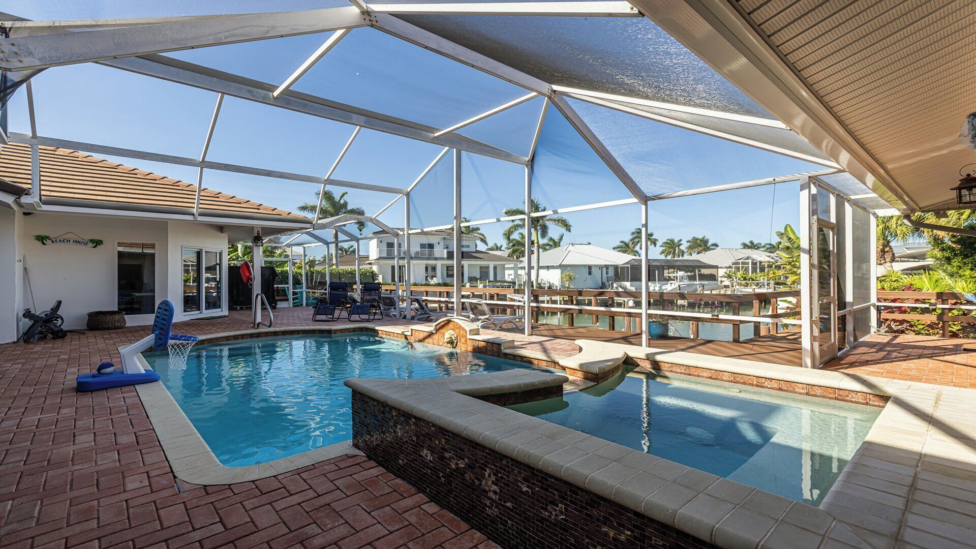 A covered swimming pool with a brick deck, lounge chairs, and a view of the surrounding area including a beach house and palm trees.