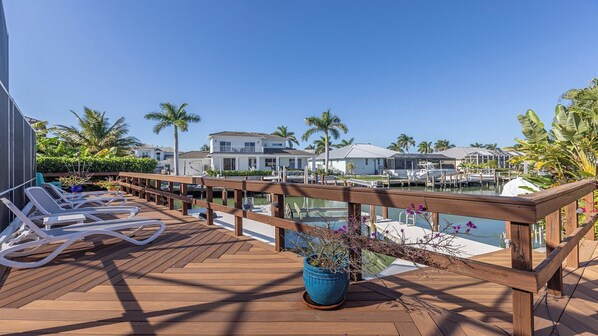 A wooden deck with lounge chairs, a wooden railing, and a view of a marina with boats and houses in the background.