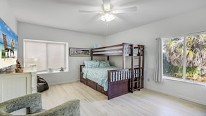 A bedroom with a bunk bed, a desk with a lamp, a chair, a ceiling fan, and a large window with a view of trees.