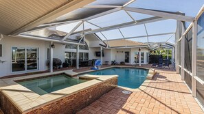 A covered outdoor swimming pool area with two hot tubs, a barbecue, and a patio with chairs and tables.