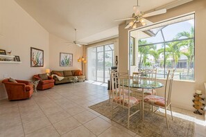 A spacious living area with a glass dining table, a ceiling fan, and a large window with a view of palm trees.