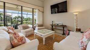 A spacious living room with a large window, a glass coffee table, and a flat-screen TV mounted on the wall.