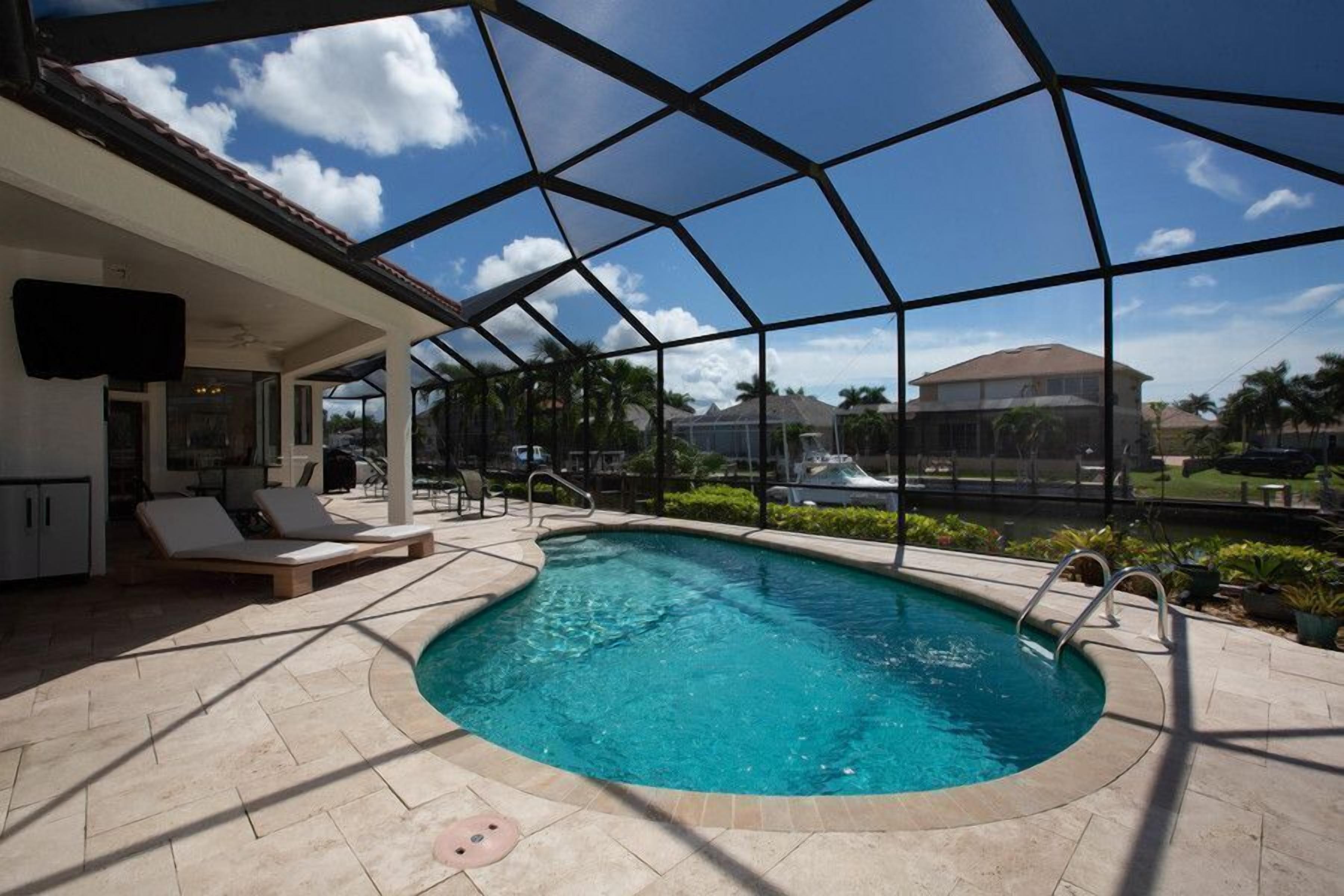 A pool area with a clear view of the sky, a covered patio with lounge chairs, and a house in the background.