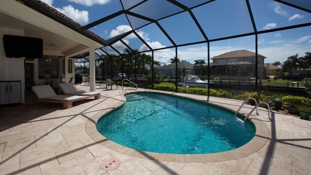 A pool area with a clear view of the sky, a covered patio with lounge chairs, and a house in the background.