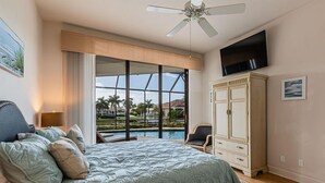 A bedroom with a bed, a ceiling fan, a TV, a large window, and a view of a pool.